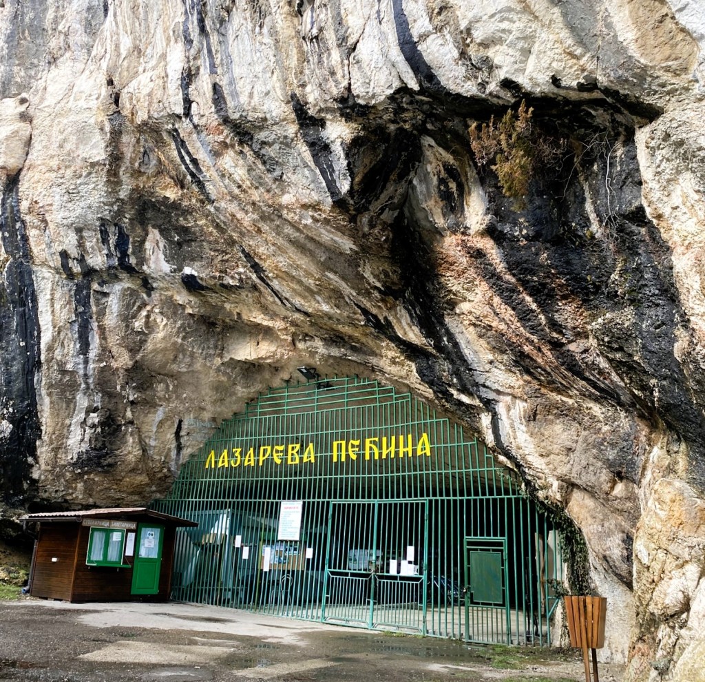 Entrance of Lazareva Cave, with a metal gate and sign in Cyrillic, surrounded by rocky terrain.