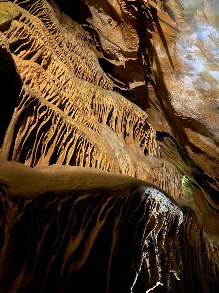 An intricate formation of rock and mineral textures inside a cave, displaying layered, striated patterns with warm lighting highlighting the contours.