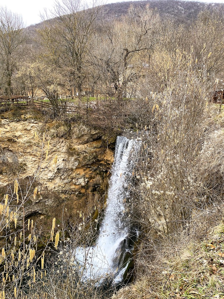 A beautiful waterfall cascading down a rocky cliff, surrounded by bare trees and shrubs during the early spring season.