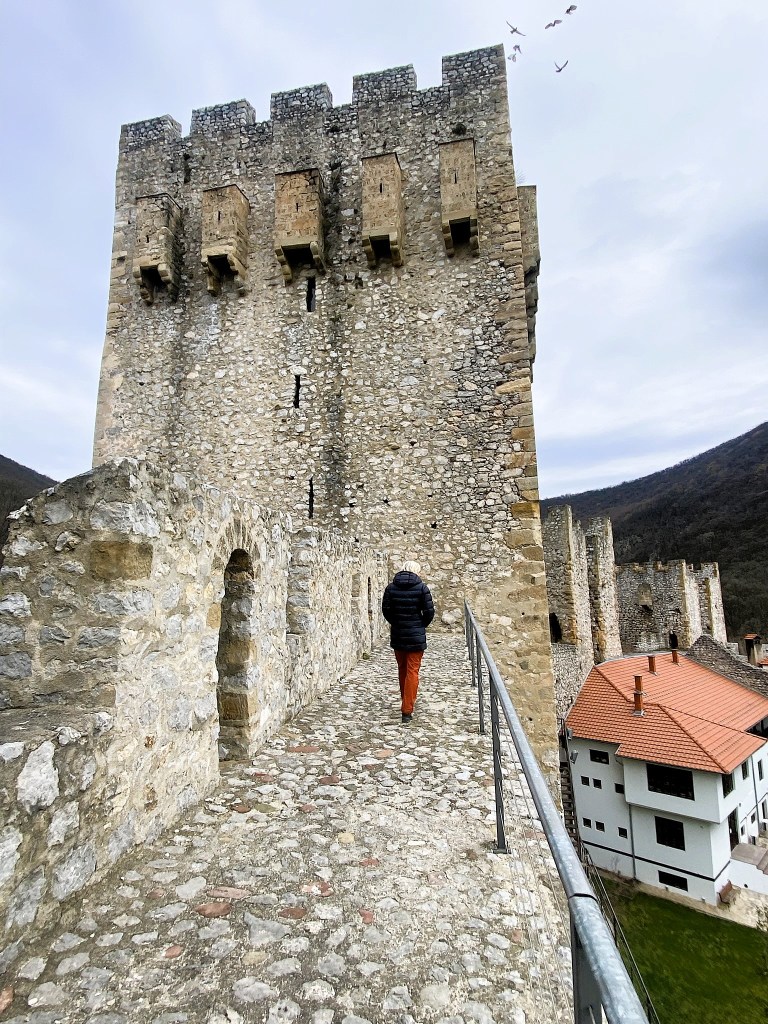 A person in a dark jacket walks along a stone pathway on top of a castle wall, with towers made of stone in the background and a house with a red roof visible nearby.
