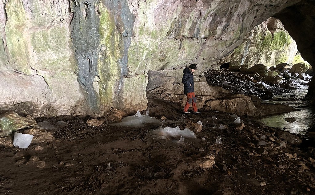 A person standing in a cave with rocky walls, examining ice formations on the ground, and surrounded by a natural and rugged landscape.