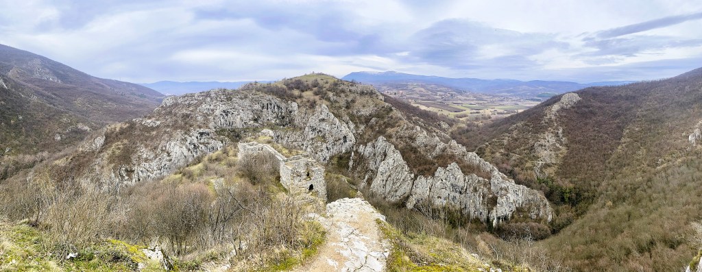 A panoramic view of a mountainous landscape featuring rocky formations and sparse vegetation, with rolling hills and valleys visible in the distance under a cloudy sky.