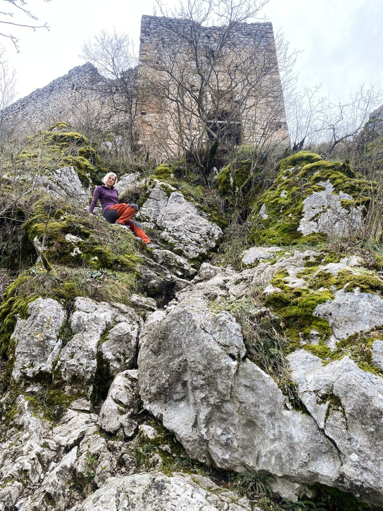 A person sitting on rocky terrain covered with moss, in front of a partially ruined stone building surrounded by sparse trees.
