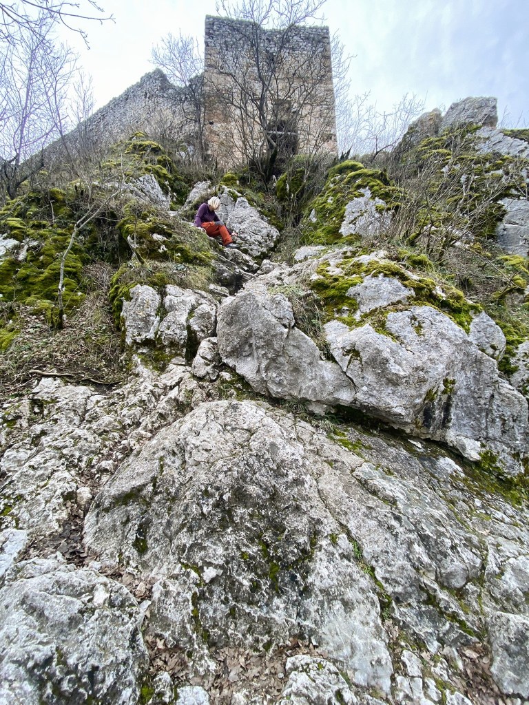 A person in orange trousers sits on rocky terrain covered with moss, with a stone structure in the background against a cloudy sky.