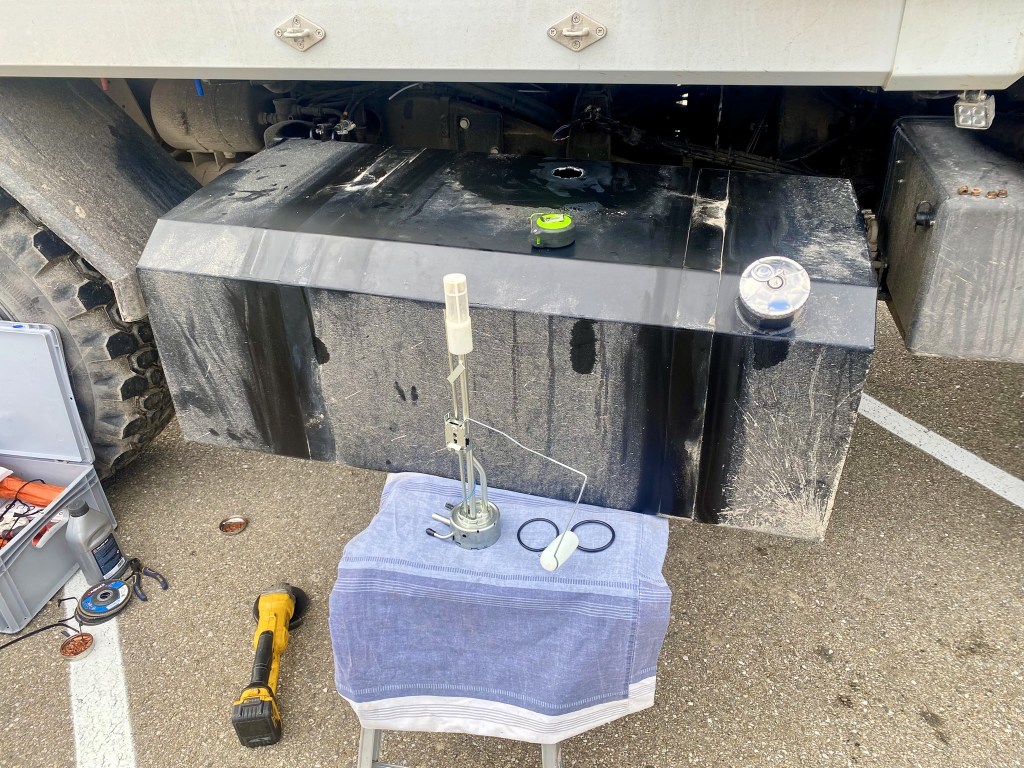 A close-up view of a black fuel tank under a truck, featuring a measuring device and tools on a towel nearby. The ground is paved, showing tire treads and additional tools in the background.