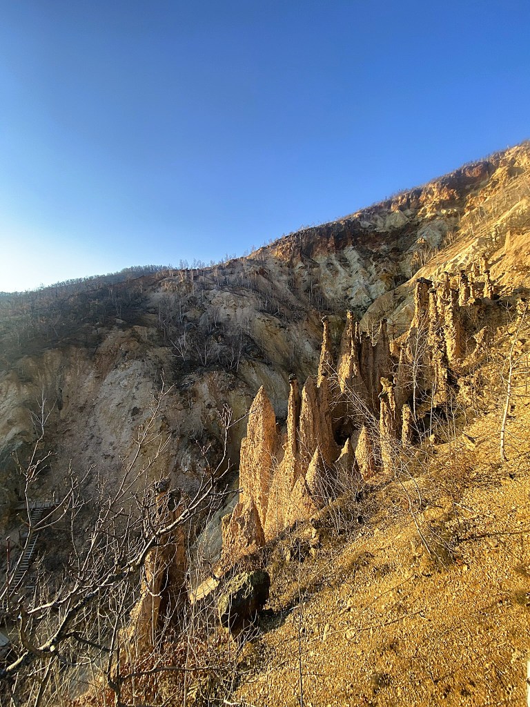 Rock formations resembling spires on a hillside, with a clear blue sky above and dry vegetation around.