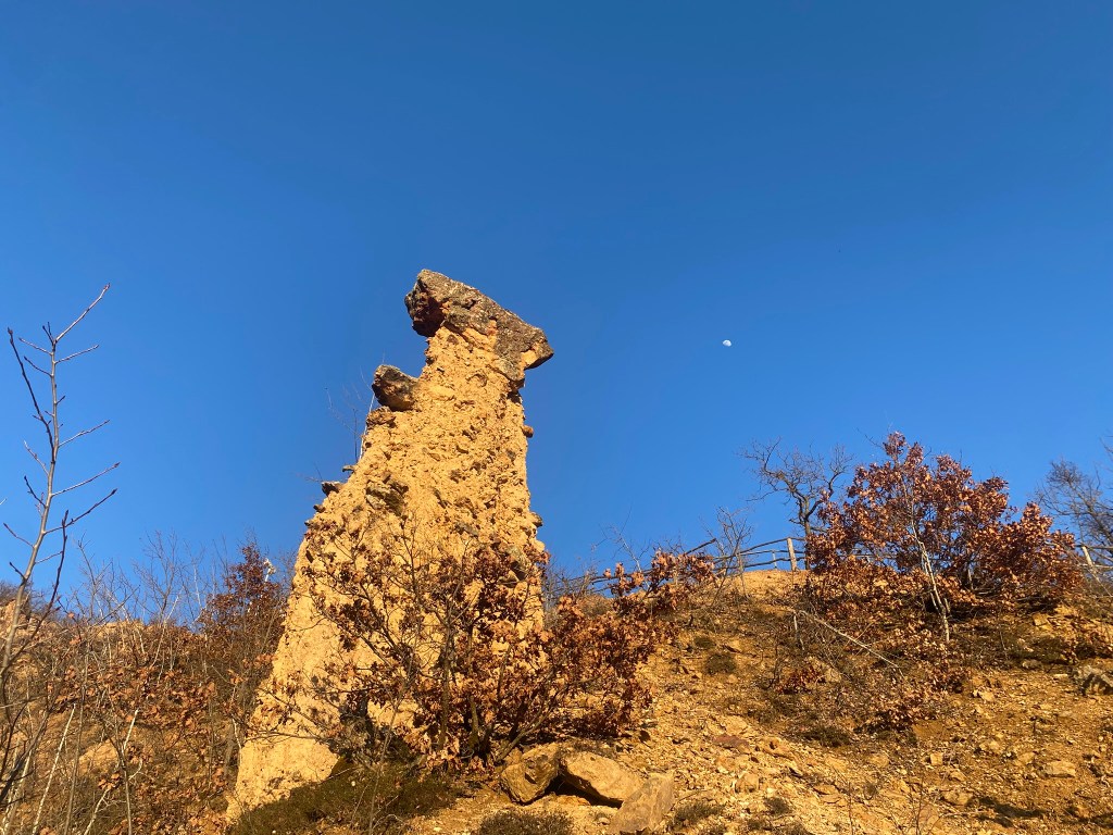 A tall, weathered rock formation standing on a hillside, with sparse vegetation and a clear blue sky. The moon is visible in the sky above.