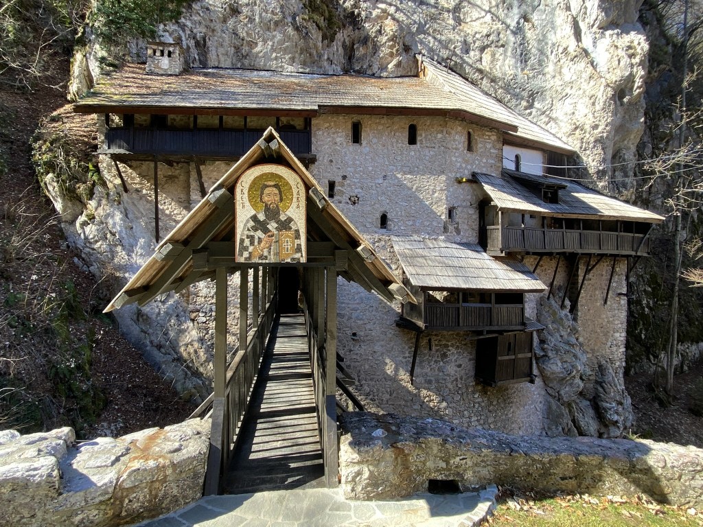 A mountain monastery with a wooden walkway leading to its entrance, surrounded by rocky outcrops and trees.