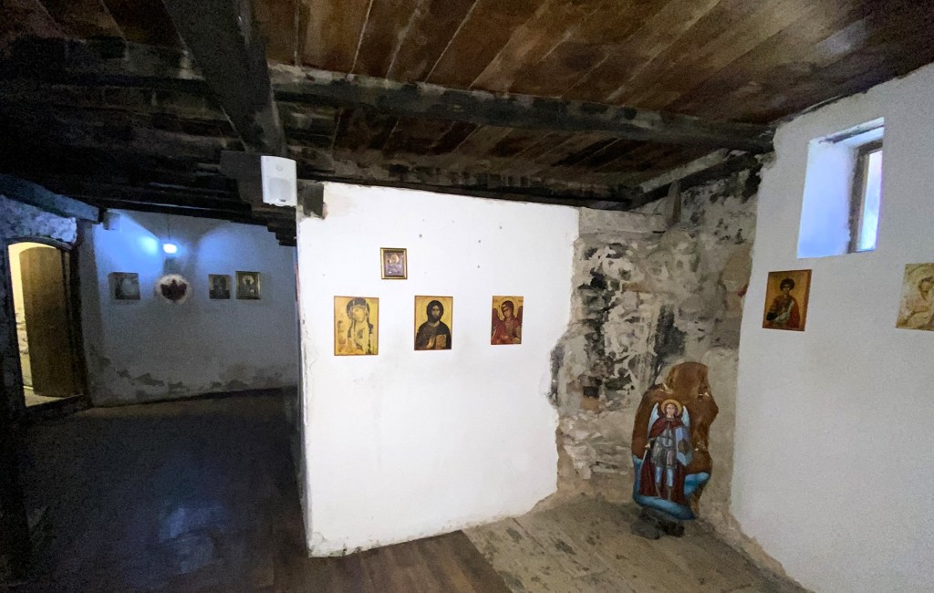Interior of a dimly lit room with wooden beams and stone walls, featuring several religious icons displayed on the walls.