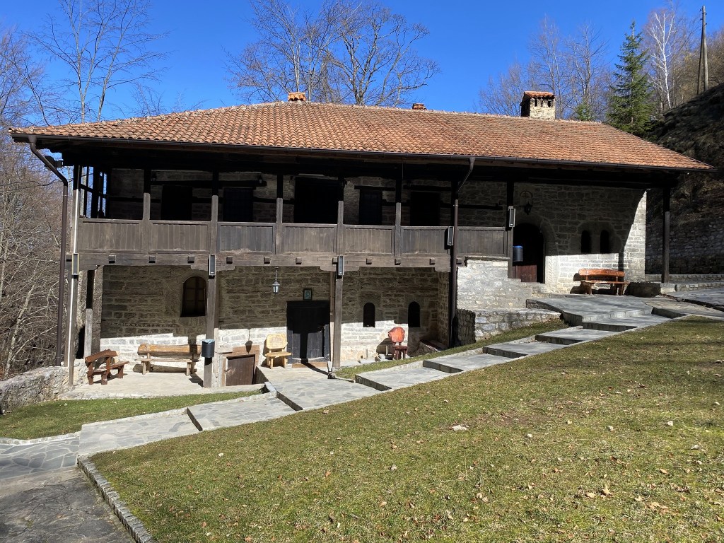 A traditional stone building with a wooden balcony, set in a natural landscape with trees and a clear blue sky.