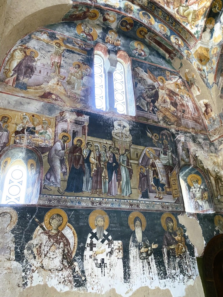 A view of the interior wall of a church featuring religious frescoes depicting scenes from the New Testament, including the crucifixion of Jesus and images of saints, with ornate details and a window allowing light to enter.