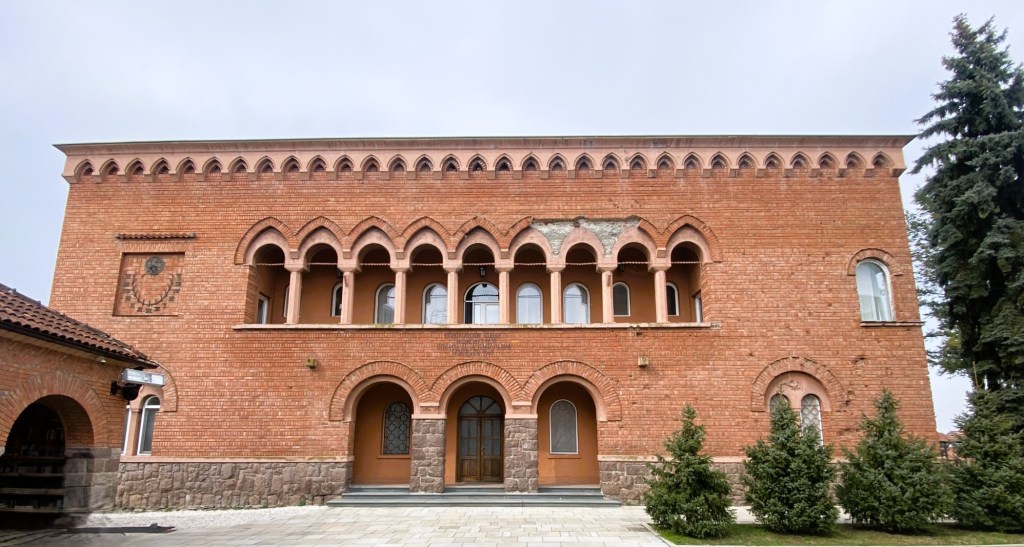 Front view of a historic red brick building featuring arched windows and doors, with decorative elements and a landscaped area.