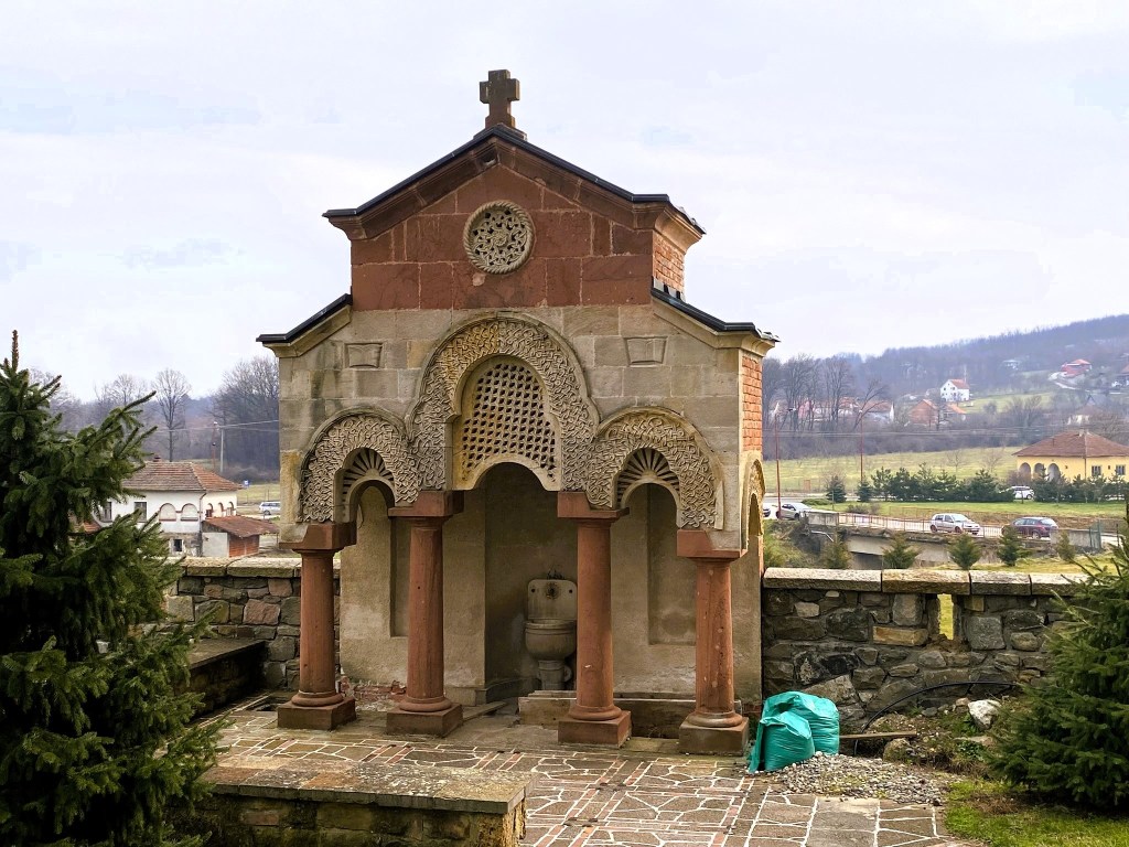 An ancient stone chapel with a decorative façade, featuring arches and columns, set against a mountainous backdrop.