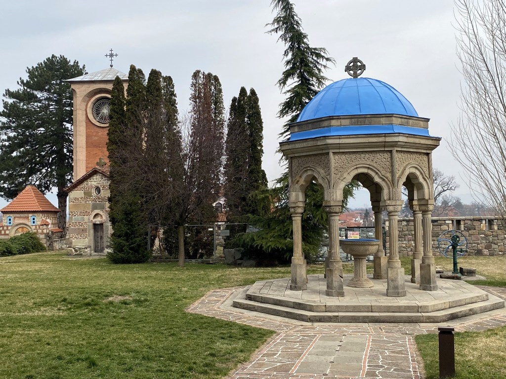 A view of a landscaped garden featuring a domed, stone gazebo with a blue roof, surrounded by tall trees and buildings with a church tower in the background.
