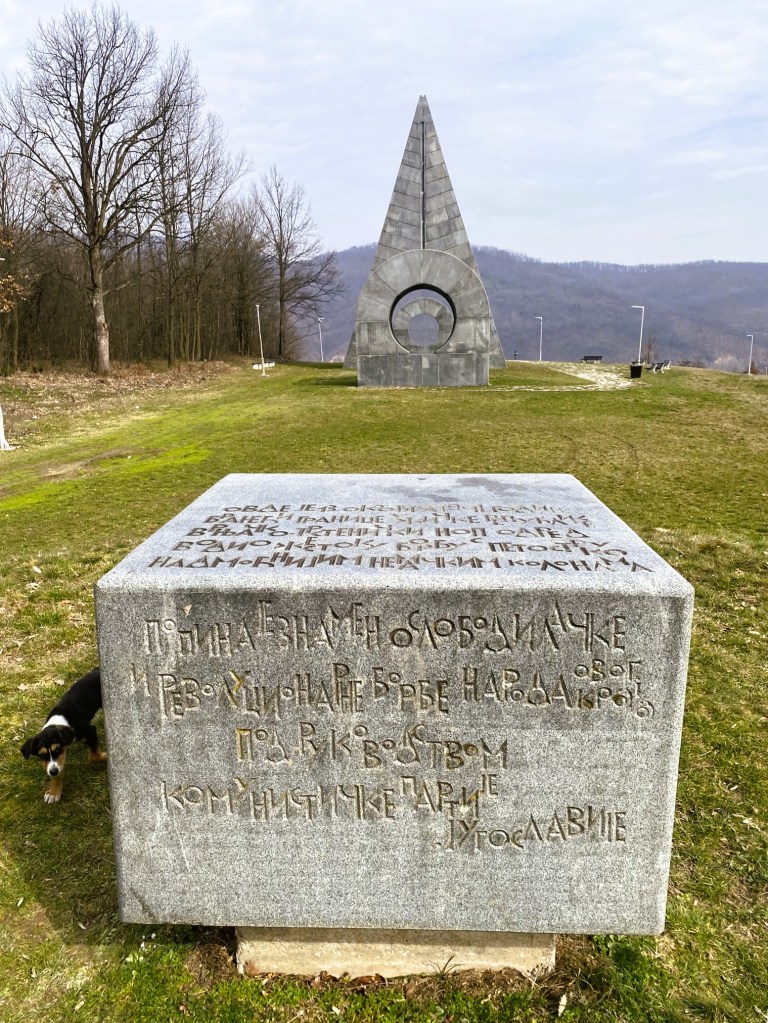 A stone monument with inscribed text and a large triangular structure in the background, surrounded by trees and grass.