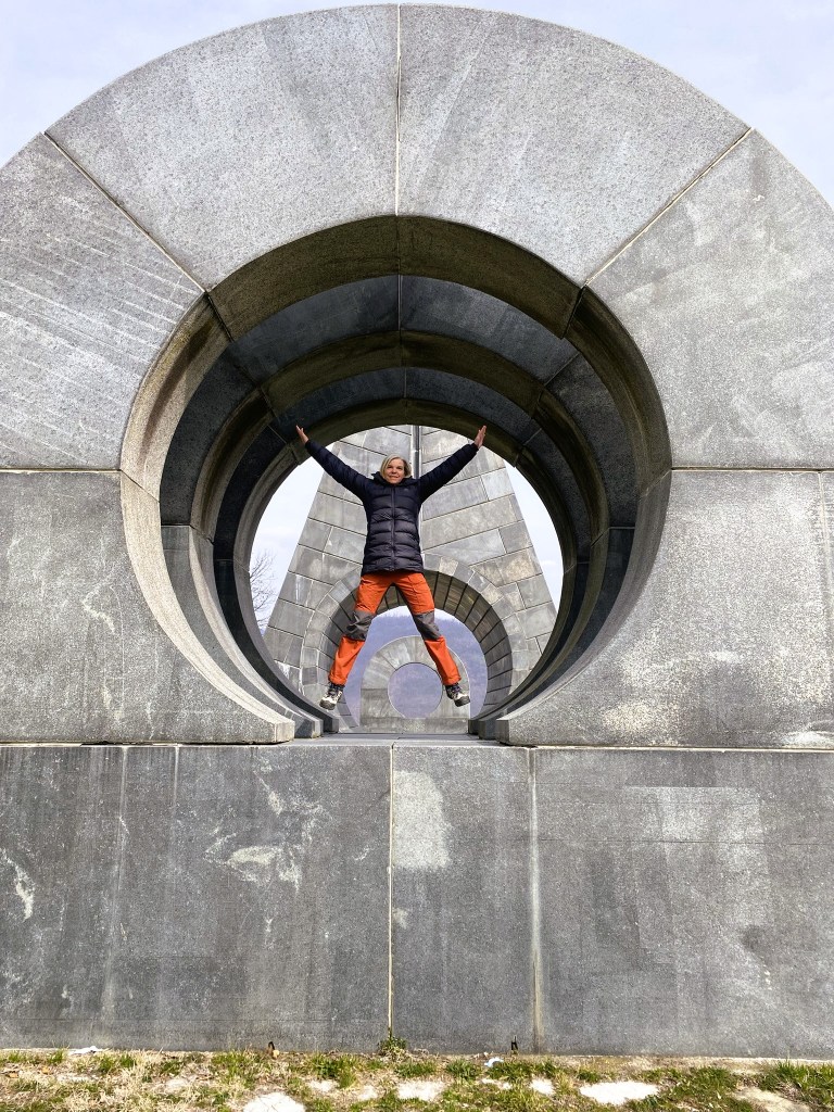 A person stands joyfully in a sculptural stone archway, with arms raised, set against a backdrop of concentric circular stone structures.