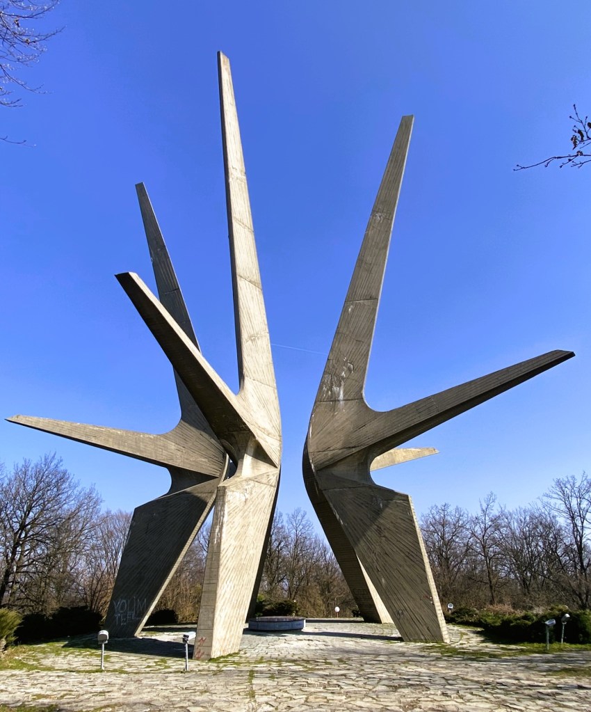 A tall concrete monument with five elongated, angular structures reaching upwards against a clear blue sky, surrounded by trees and greenery.