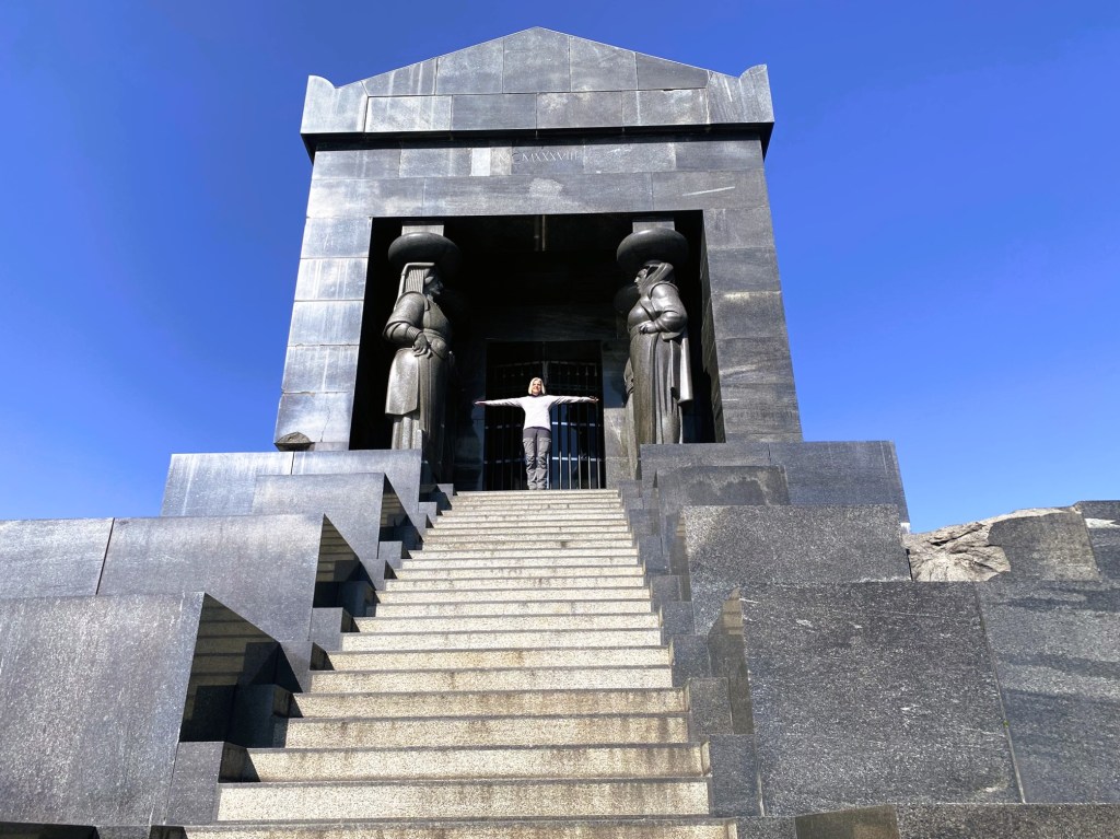 A person standing on steps in front of a large stone monument, flanked by two statues, under a clear blue sky.