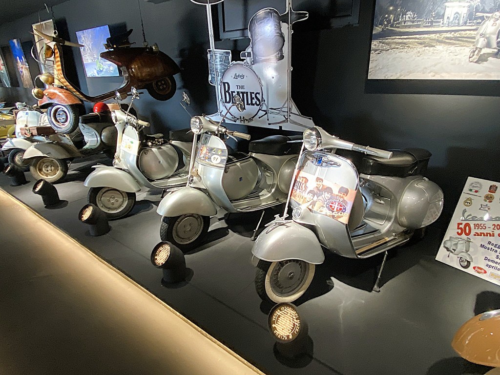 A display of vintage silver scooters lined up in a darkened space, featuring decorative elements and a prominent 'The Beatles' theme.