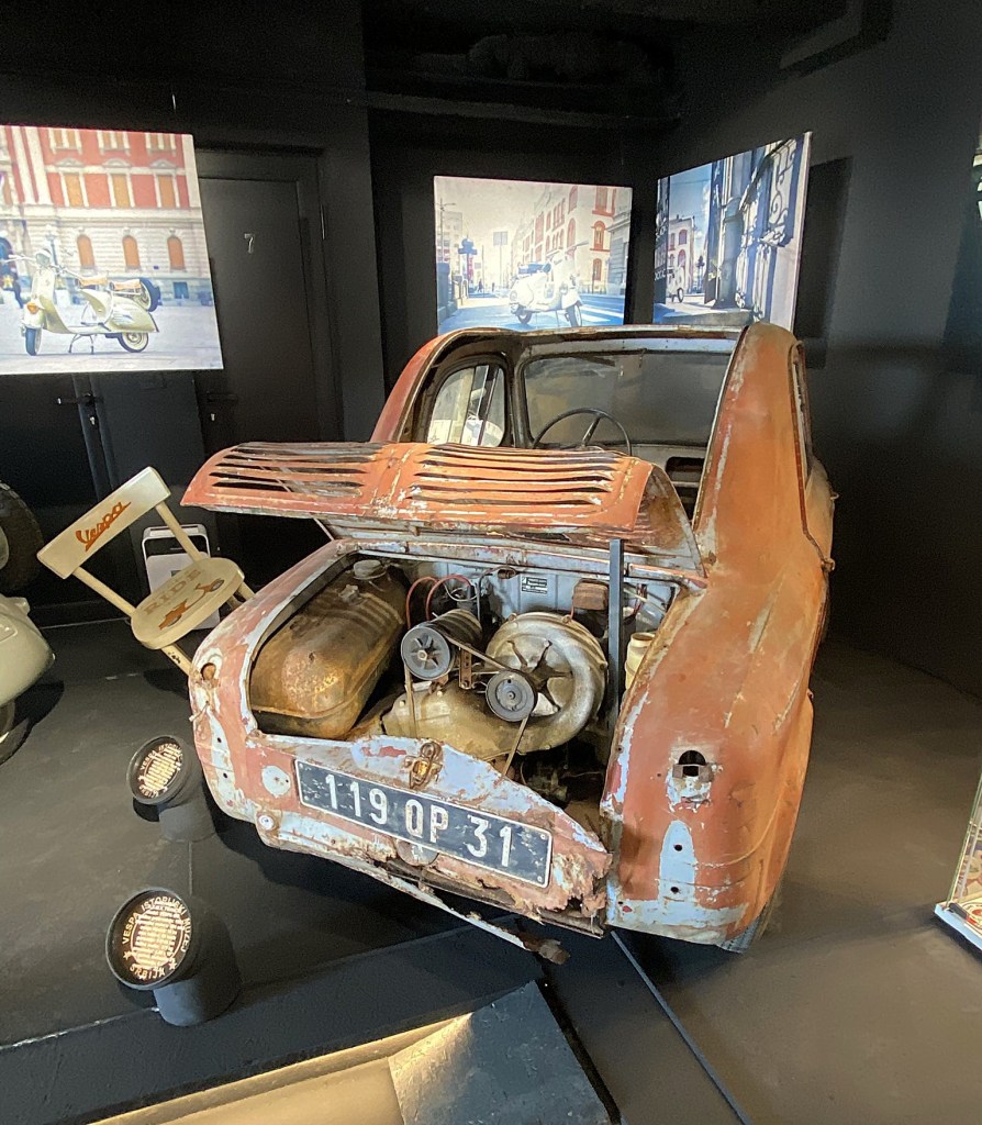 A vintage car with a rusty exterior on display, featuring an open engine compartment, set against a dark background with artwork depicting scooters.