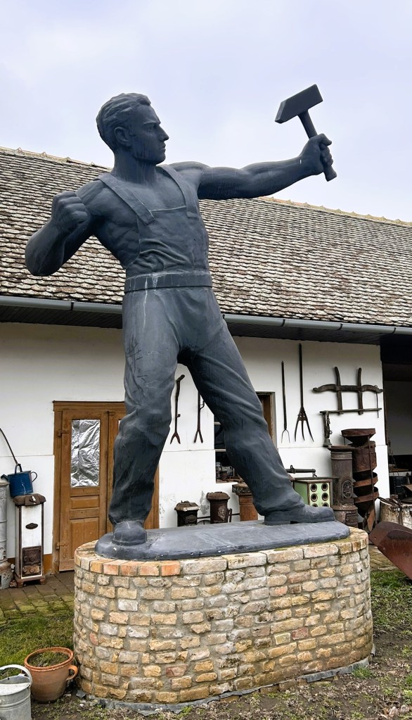 A large black statue of a muscular man holding a hammer, standing on a brick pedestal in front of a building with various tools and items at the sides.