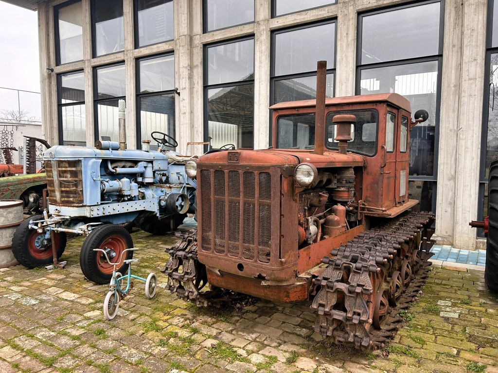 An antique tracked tractor with a rusty red finish, parked next to a blue tractor, in front of large glass windows.