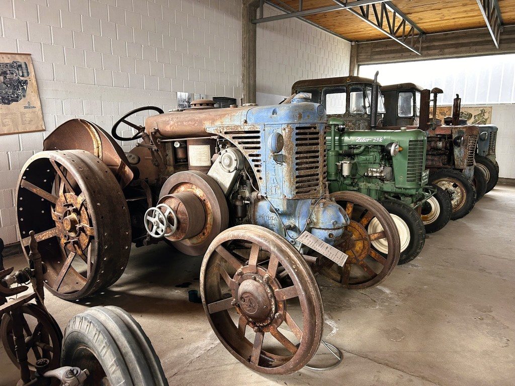 A collection of vintage tractors displayed indoors, featuring a blue tractor at the forefront with large spoked wheels, and several other tractors in a row behind it, showcasing different colours and designs.