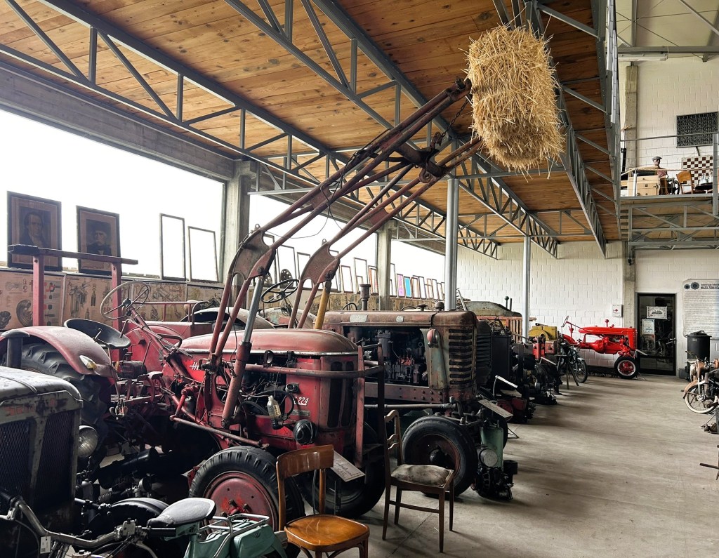 A collection of vintage tractors and agricultural machinery inside a spacious warehouse with wooden beams and framed pictures on the walls.