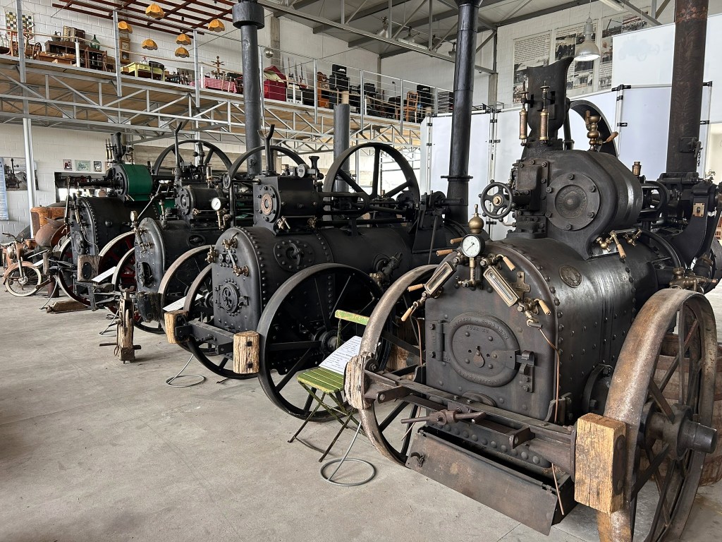 A row of vintage steam engines displayed in an industrial space, showcasing intricate machinery and wheels.