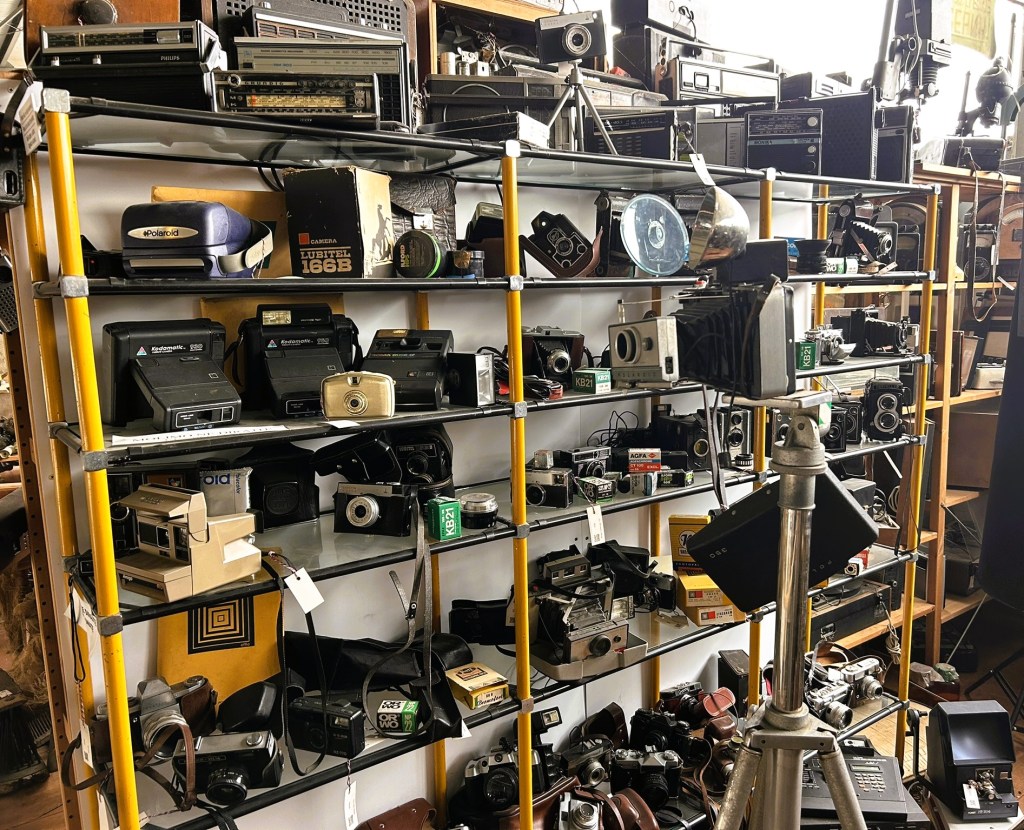 A display of vintage cameras and audio equipment on shelves, featuring various models and accessories, with a camera on a tripod in the foreground.