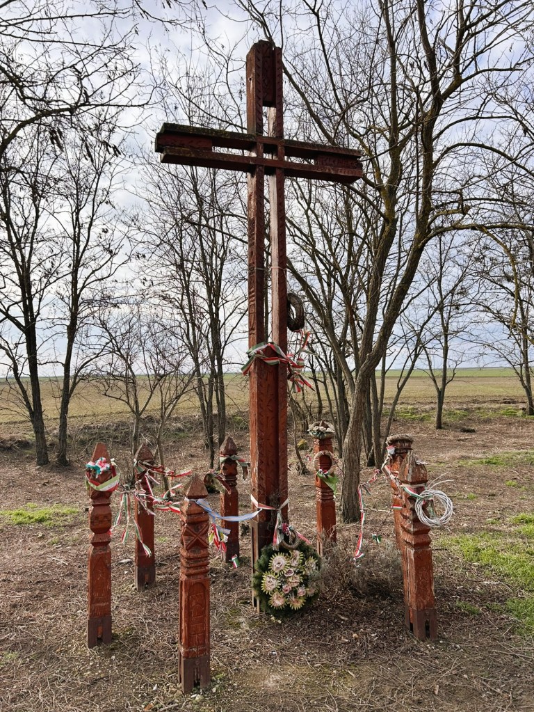 A wooden cross surrounded by carved wooden posts, decorated with ribbons and a floral wreath, set in a rural landscape with bare trees in the background.