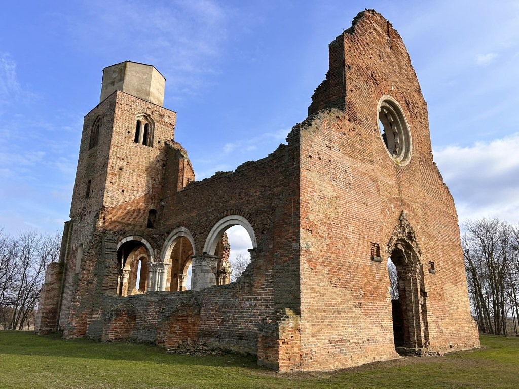 Ruins of a brick building with an arched doorway and a tower, set against a blue sky with scattered clouds.
