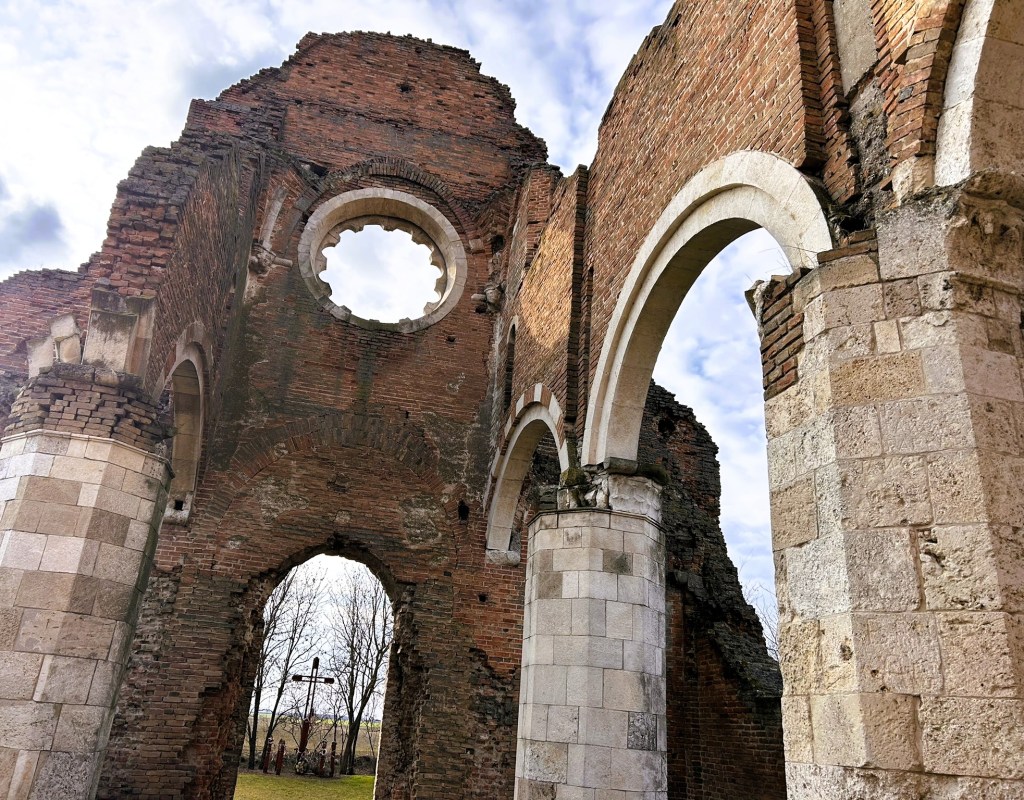 The ruins of an old brick structure featuring arches and a large circular window, set against a cloudy sky.