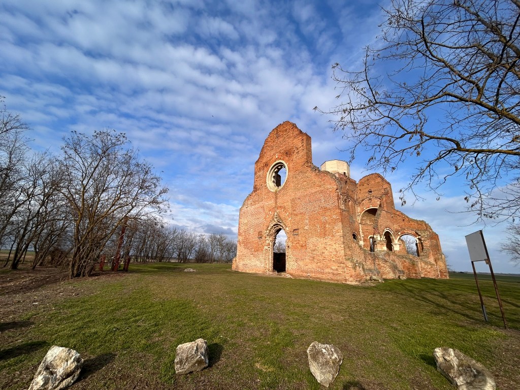 Ruins of an ancient brick building set against a cloudy sky, surrounded by bare trees and green grass.