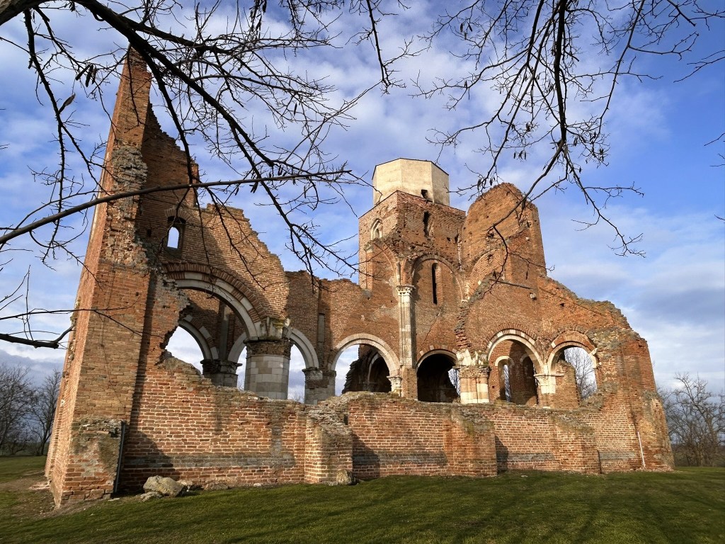 Ruined brick structure of a historical building with arched windows and a small tower, surrounded by trees and grass under a cloudy sky.