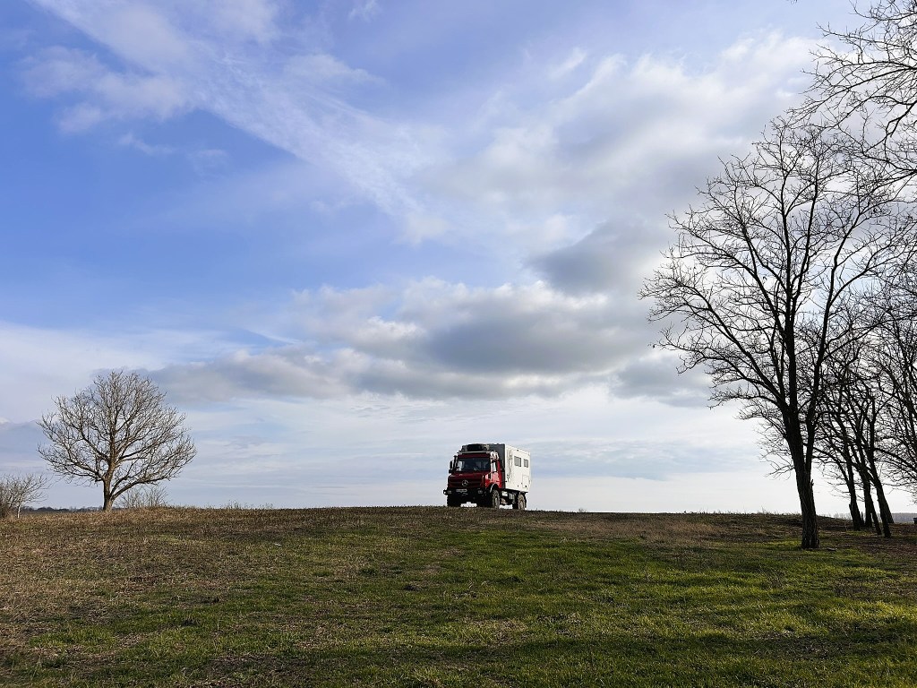 A red and white expedition vehicle parked on a grassy hill, with a bare tree and a few leafless trees in the background under a partly cloudy sky.