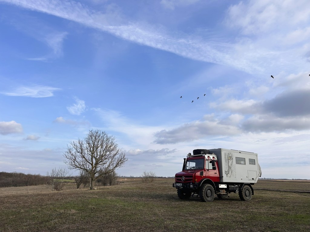 A red overland vehicle parked in an open field under a blue sky, with a few clouds and birds flying overhead. A single tree stands nearby, adding to the natural landscape.