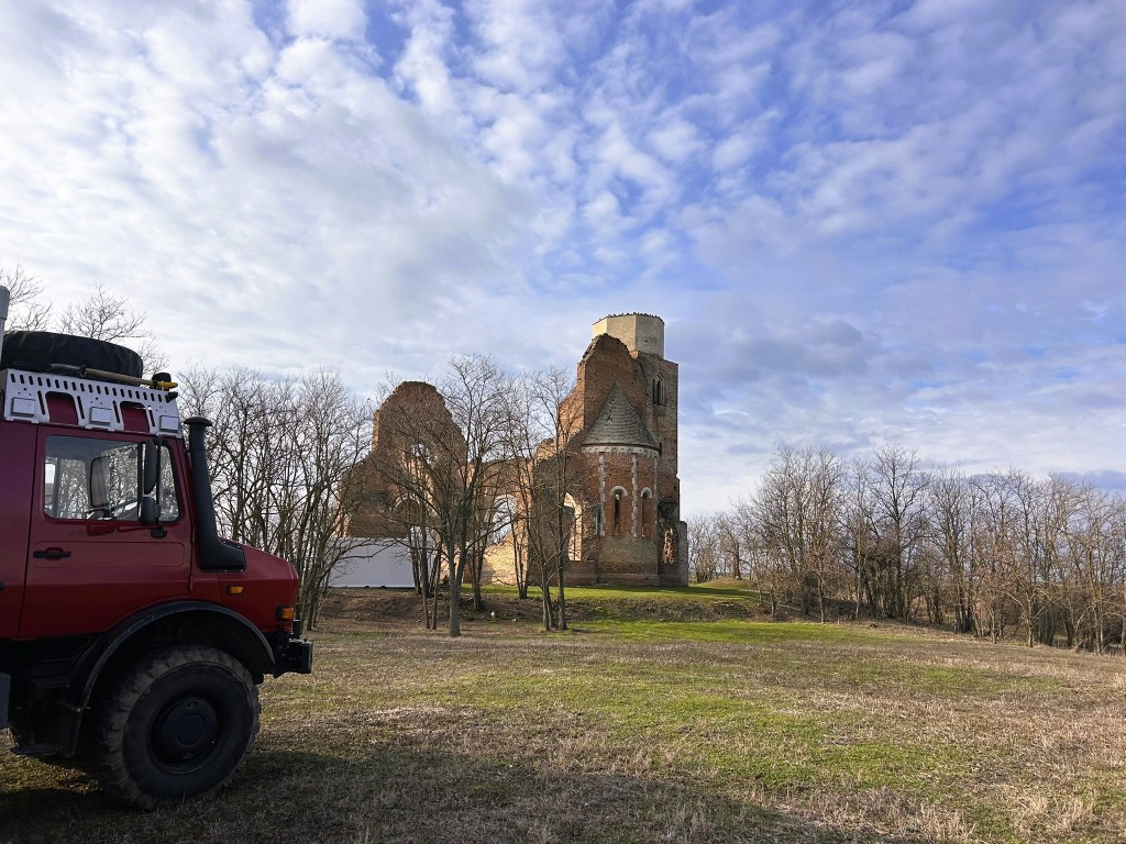 A red off-road vehicle parked in a grassy area, with a partially ruined brick church and a cloudy sky in the background.