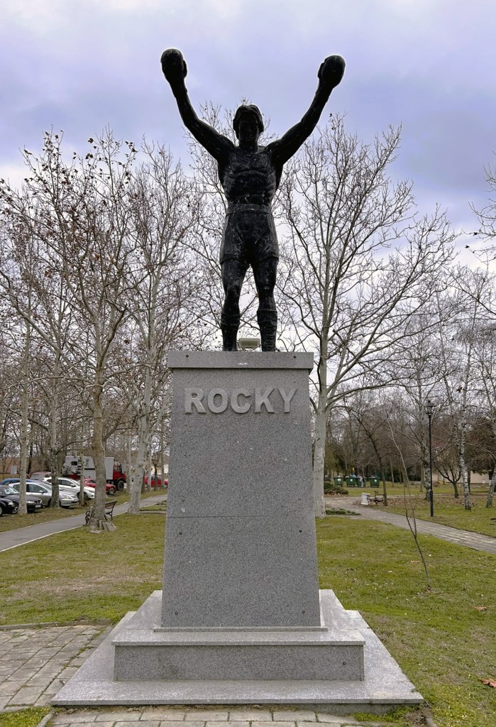 Statue of Rocky Balboa with arms raised in triumph, positioned on a stone pedestal engraved with 'ROCKY', set in a park with bare trees.
