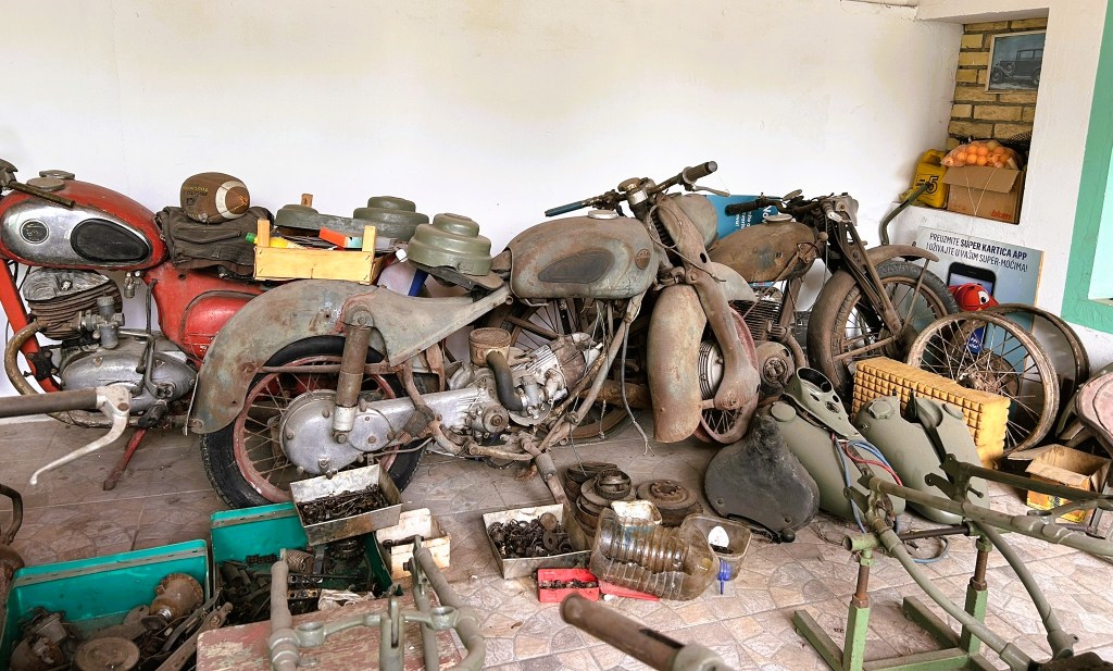 A collection of vintage motorcycles in an old garage, featuring two rusty bikes with battered frames and a variety of mechanical parts and tools scattered around.