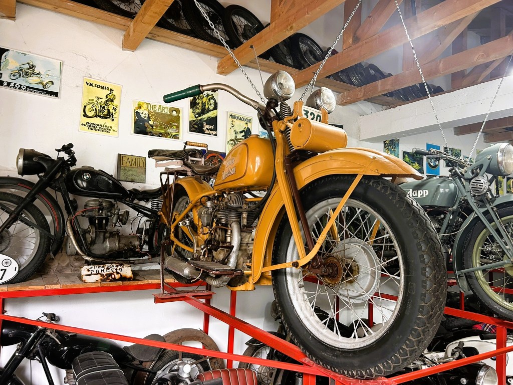 A vintage yellow motorcycle displayed on a red stand, surrounded by other classic bikes and framed vintage motorcycle advertisements on the walls.
