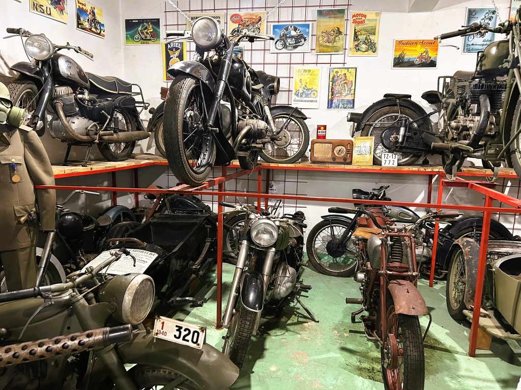 A collection of vintage motorcycles displayed in a small museum setting. The image features several motorcycles of varying styles and ages, stacked on red shelving. Vintage posters are hung on the wall, and some old items like a radio and memorabilia are visible among the motorcycles.