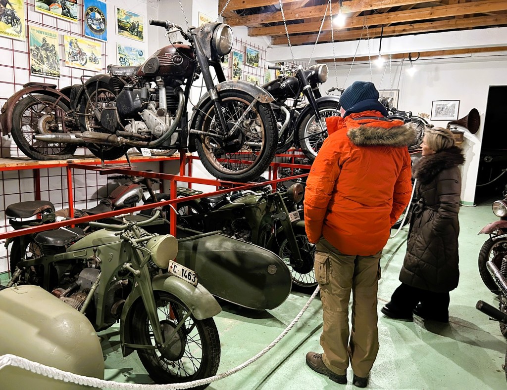 A vintage motorcycle display in a museum with various models on shelves, including a classic motorcycle on the top shelf. Two visitors, one in an orange coat and another in a brown coat, observe the exhibits.
