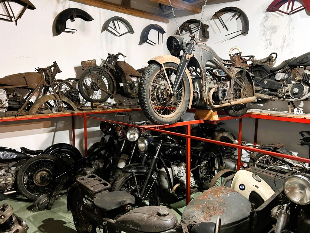 A cluttered workshop filled with vintage motorcycles and motorcycle parts displayed on shelves, showcasing various models and types in varying states of restoration.