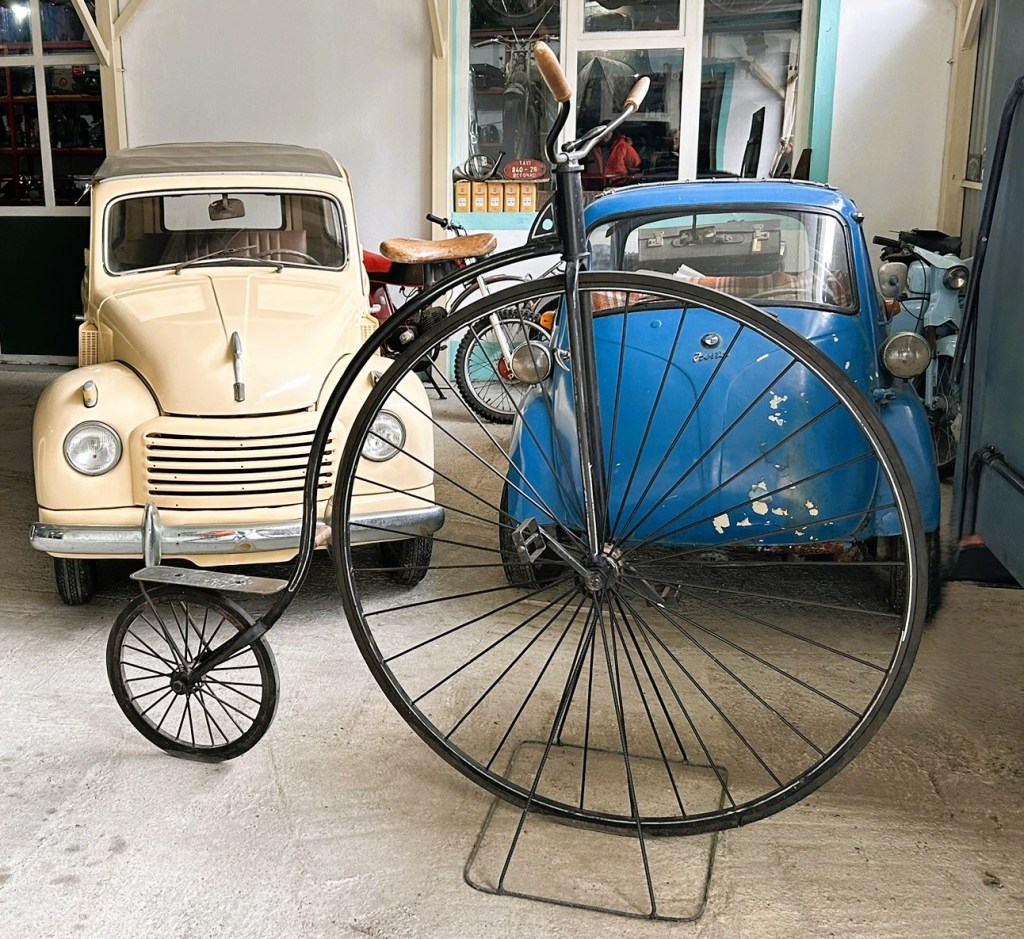 A vintage penny-farthing bicycle stands in the foreground, with a cream-coloured classic car and a blue car in the background, set in an indoor garage space.