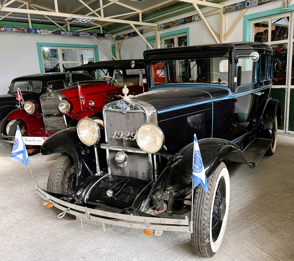 A vintage 1929 black car with blue accents, displayed in a garage alongside other classic cars. The car features round headlights and flags mounted on the front.