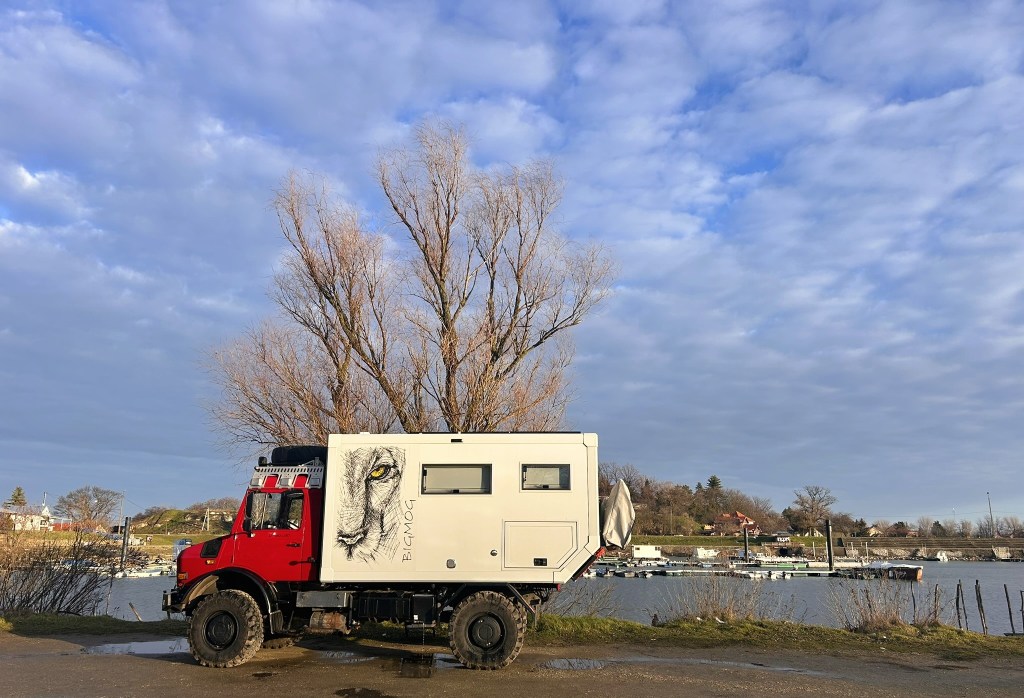 A red off-road vehicle with a camper body, featuring a lion's face illustration, parked near a lake with trees and boats in the background under a cloudy sky.