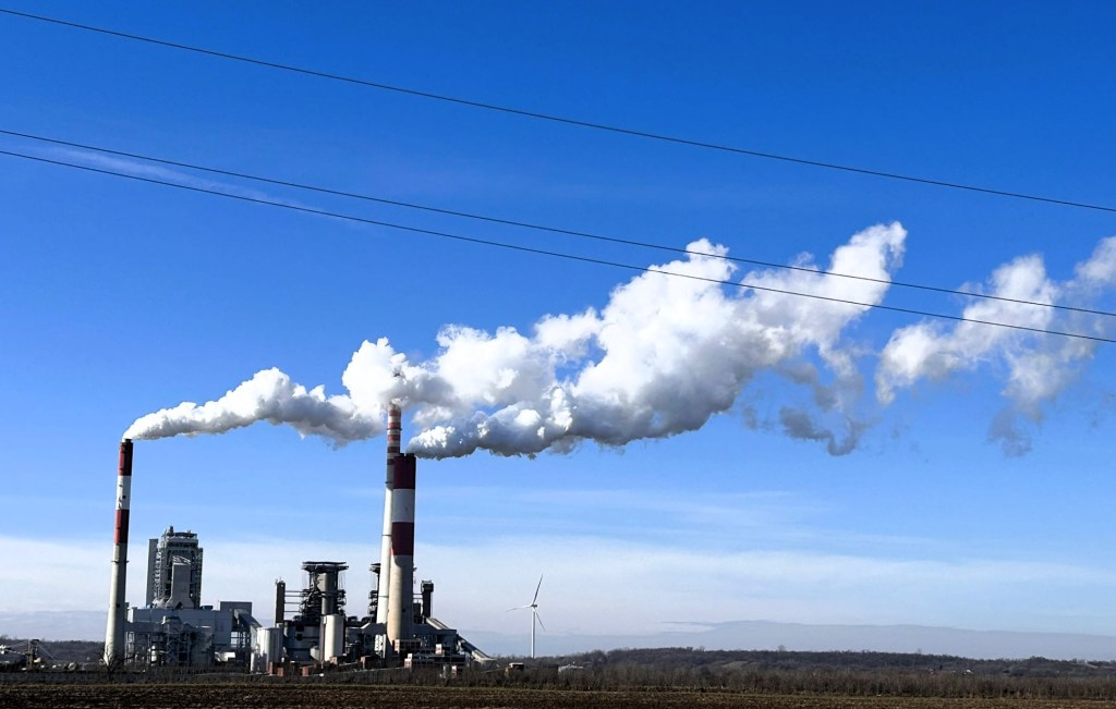 Industrial power plant with two tall smokestacks emitting white smoke against a clear blue sky, featuring wind turbines in the background.