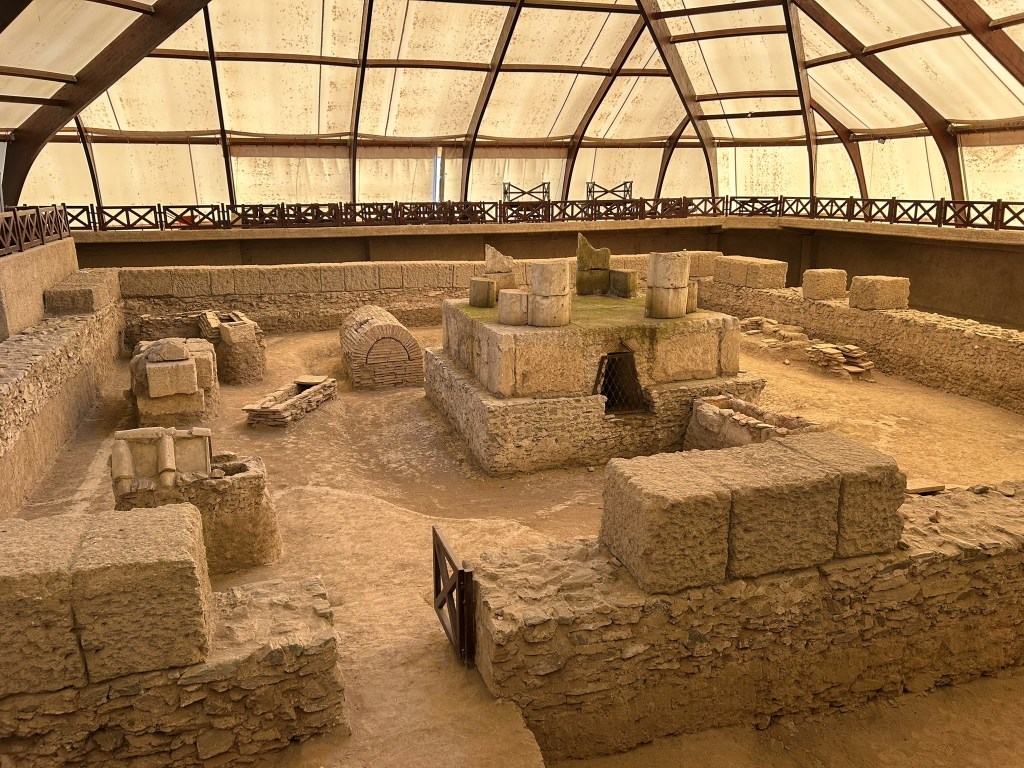 An archaeological site covered by a glass roof, featuring stone structures and ruins, with a central platform surrounded by walls and ancient artifacts.