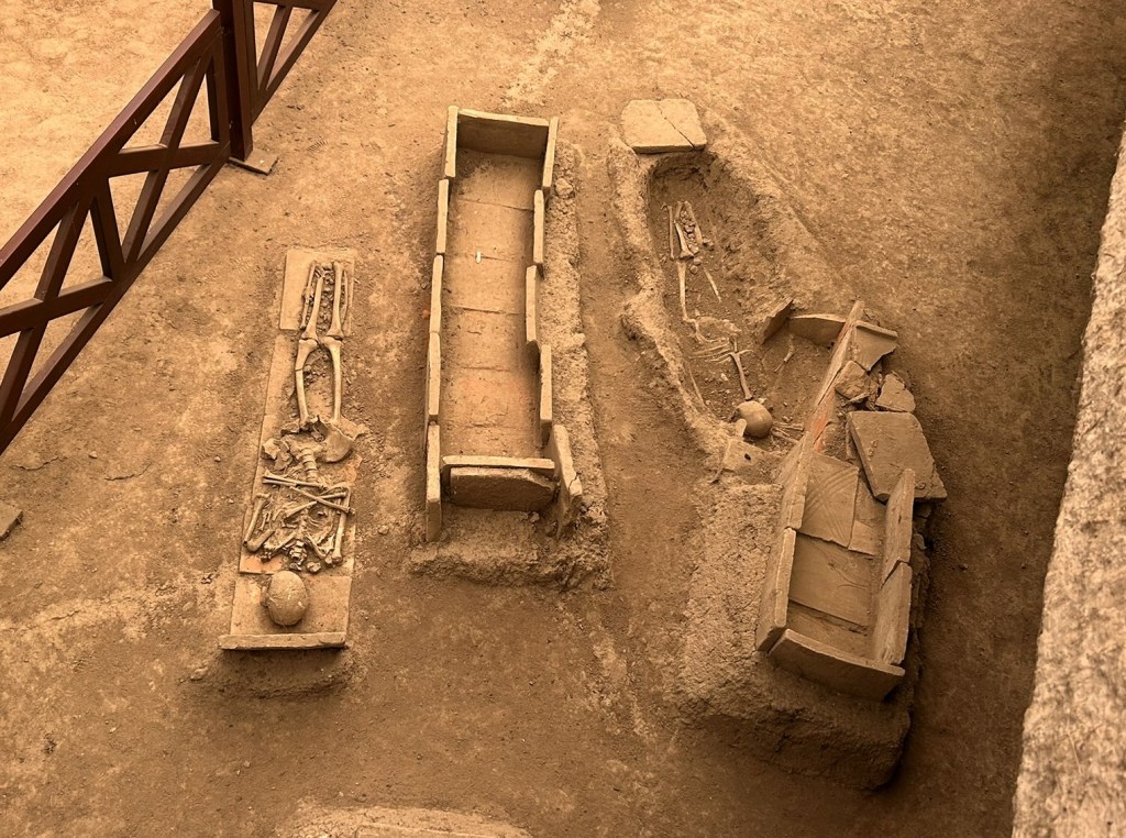Three ancient burial sites with exposed skeleton remains, located in a sandy environment, surrounded by a wooden barrier.